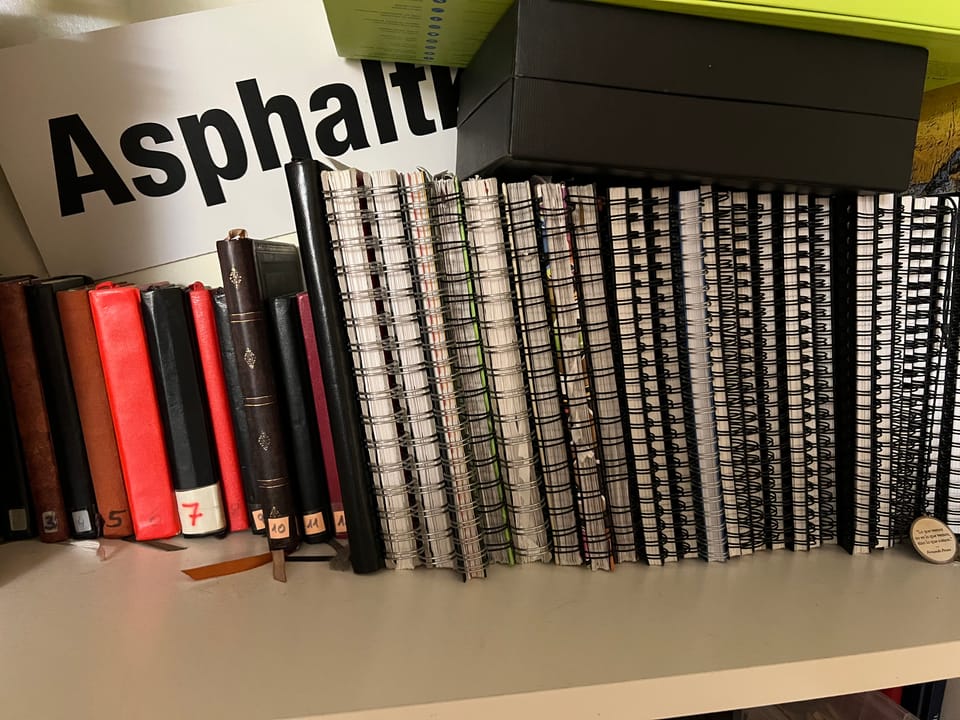 Regal mit mehreren unterschiedlich großen und gebundenen Notizbüchern und Ordnern, im Hintergrund ein Schild mit dem Schriftzug 'Asphaltkälte'